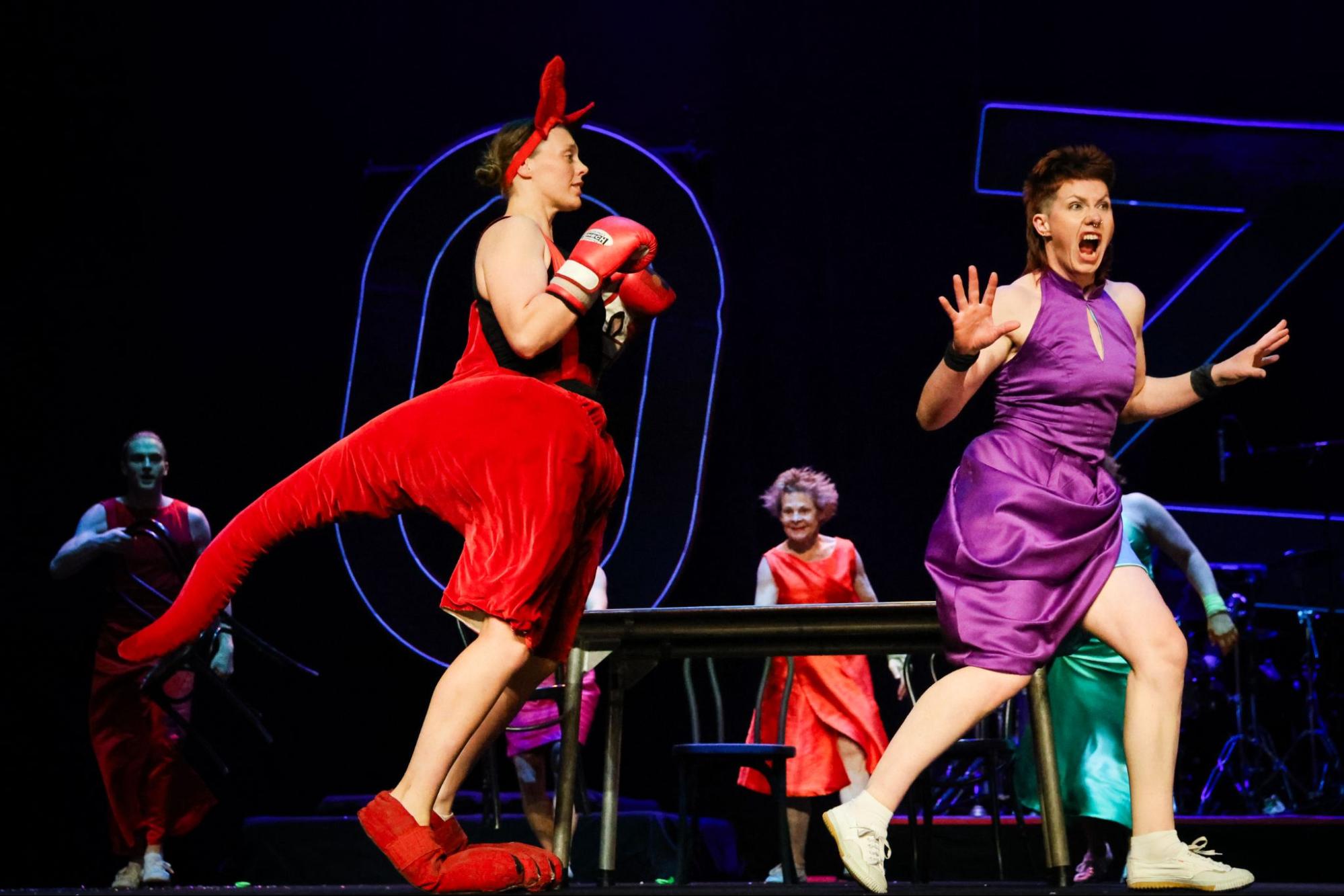 A performer dressed as a red kangaroo with red ears, big red shoes and red boxing gloves chases after a frightened performer in a purple dress.