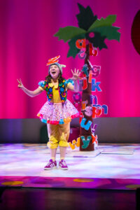 A performer in colorful clothing including a tutu and patterned vest stands in front of a coconut tree. Lowercase letters with googly eyes cling to the tree.