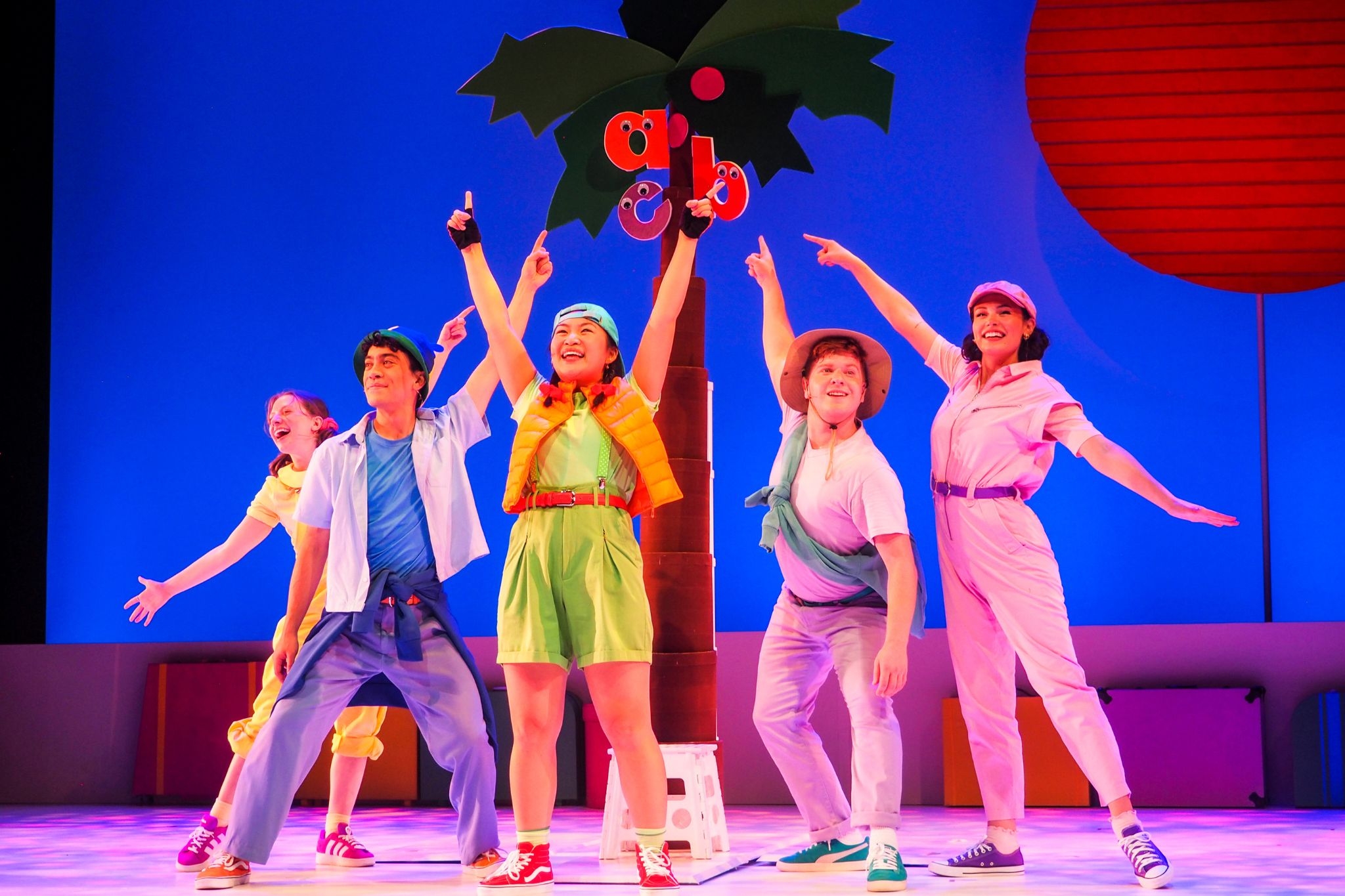 Lowercase letters "a," "b" and "c" climb to the top of the coconut tree. Five performers wearing colorful clothing smile and point up at the letters.