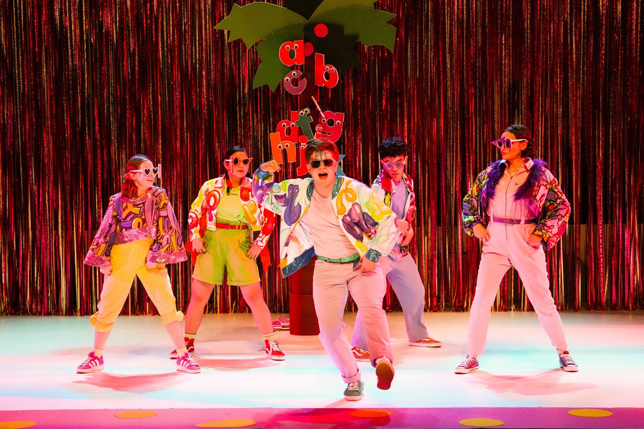 Five performers in colorful jackets and sunglasses dance in front of a coconut tree and fringe backdrop. Lowercase letters with googly eyes cling to the tree's trunk.