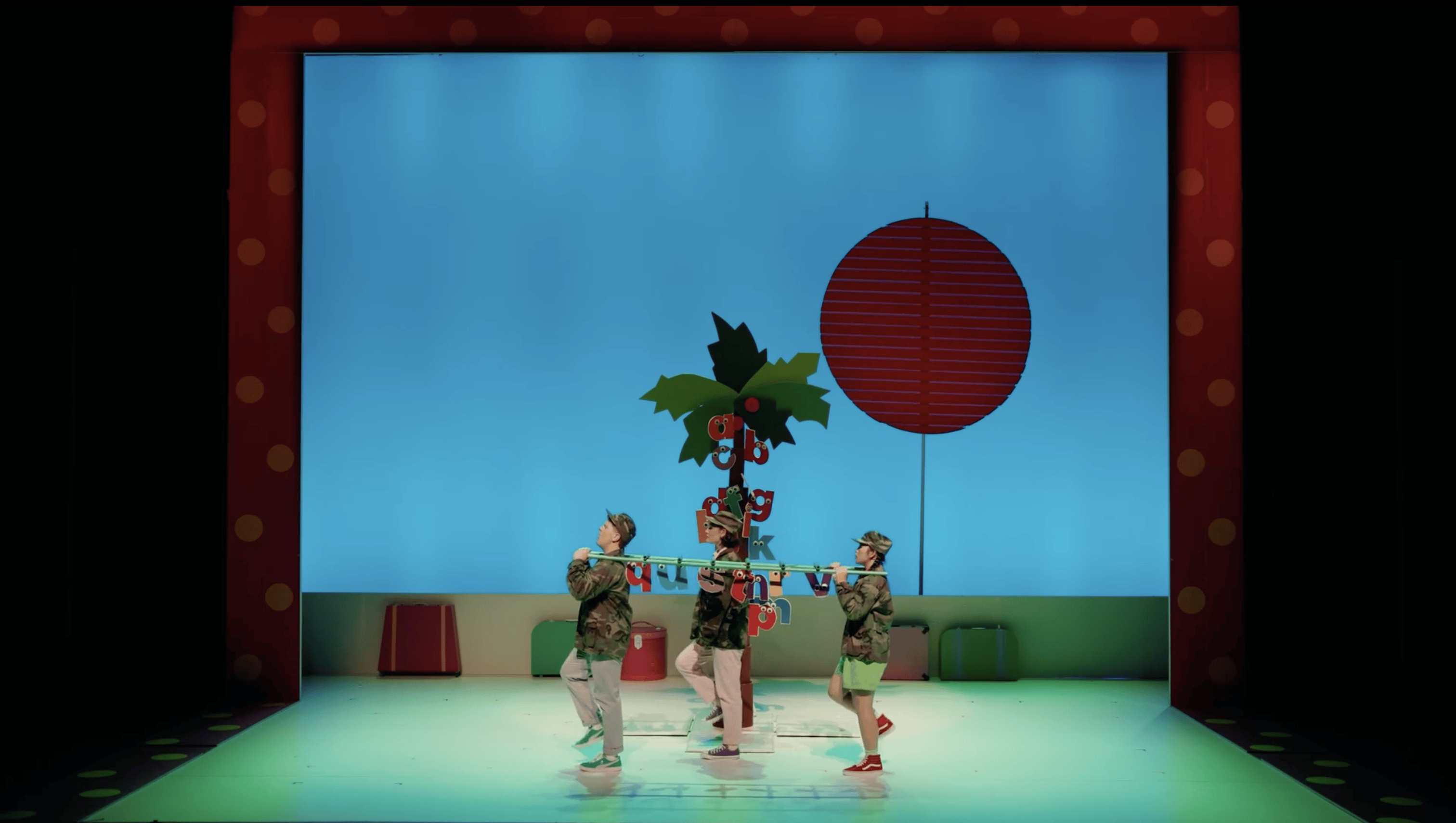 Three performers center stage with a coconut tree standing between them and a blue screen with an orange sun