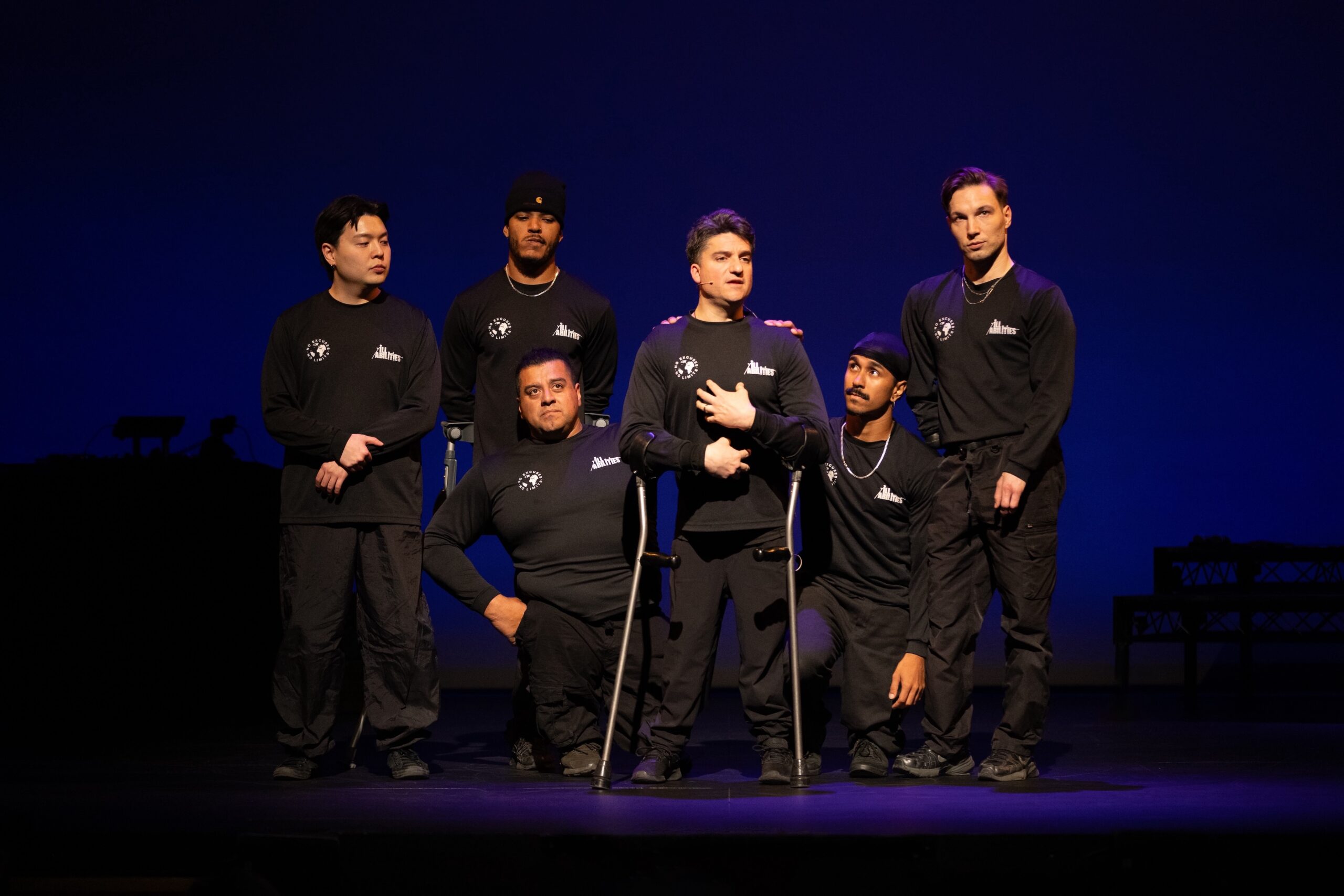Six performers with physical disabilities stand as a group while one in the center, on crutches, speaks.