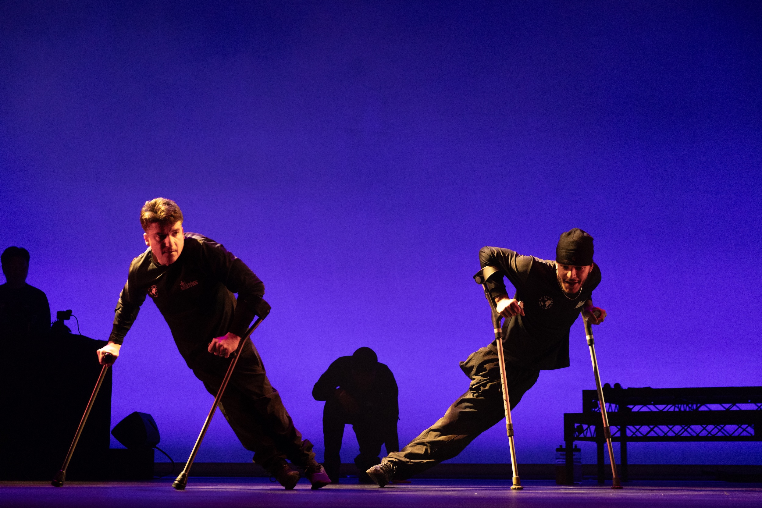 Two performers dressed in all black dance on their crutches in front of a purple background 
